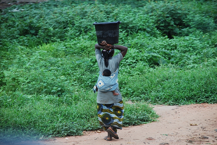 Frau mit Kind auf dem Rücken und einem Zuber auf dem Kopf